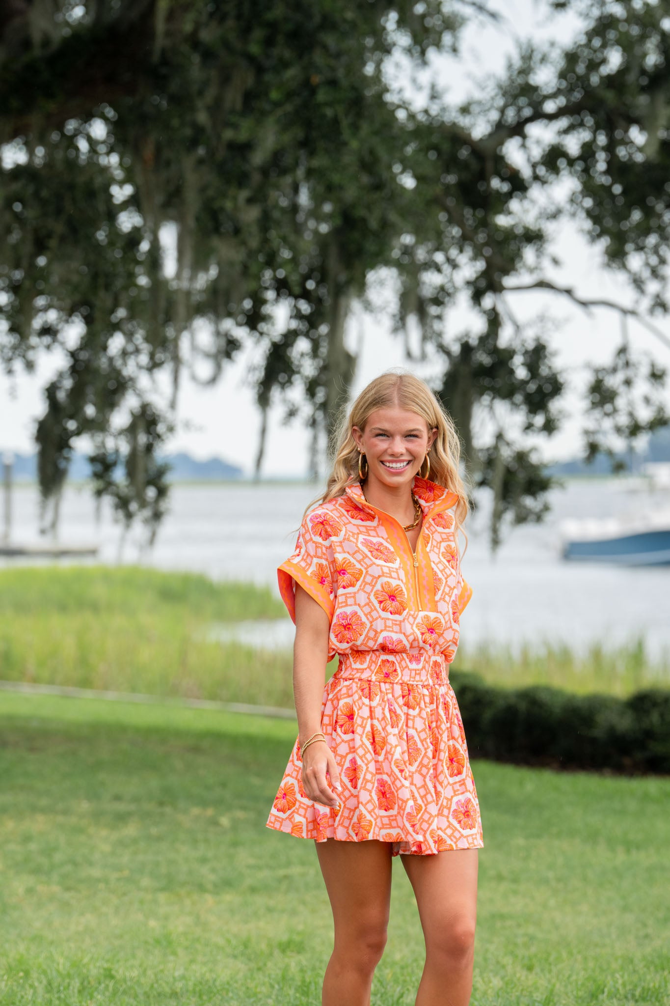 A person in a vibrant orange Poppy Pullover - Floral Crochet top smiles while standing on grass near the waterfront. A large tree with hanging moss invites joy into the serene scene in the background.