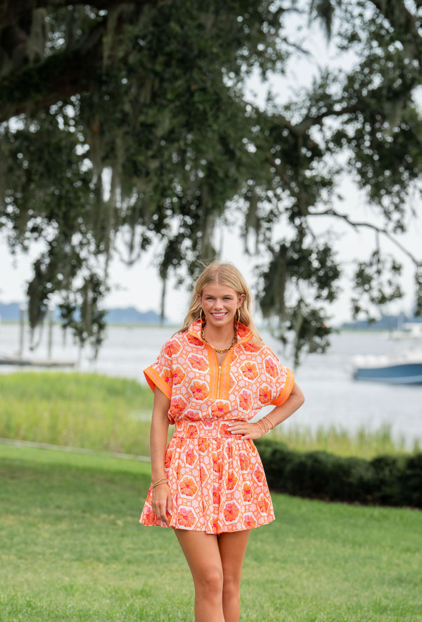 Dressed in the Swing Short - Floral Crochet from Emily McCarthy Clothing, a person stands on the grass, smiling with one hand on their hip. The scenic backdrop features a river, a dock, and large trees draped with hanging moss.