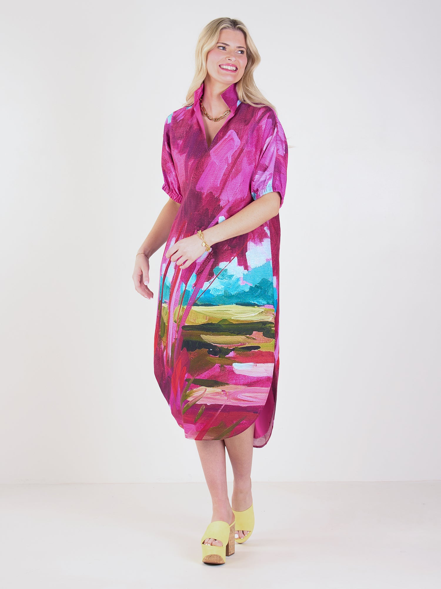 A woman with blonde hair smiles against a white background, wearing the Poppy Caftan with a hand-painted print inspired by a Lowcountry sunset and yellow heels.