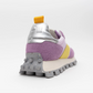 Step into the Osaka Lilac Haze Re-Nylon: a vibrant sneaker with a soft purple texture, yellow and pink accents, and a robust gray platform midsole featuring raised circular grips. The back highlights a metallic silver heel with a yellow pull tab set against a pure white background.