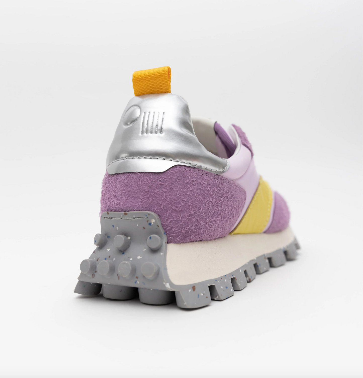 Step into the Osaka Lilac Haze Re-Nylon: a vibrant sneaker with a soft purple texture, yellow and pink accents, and a robust gray platform midsole featuring raised circular grips. The back highlights a metallic silver heel with a yellow pull tab set against a pure white background.