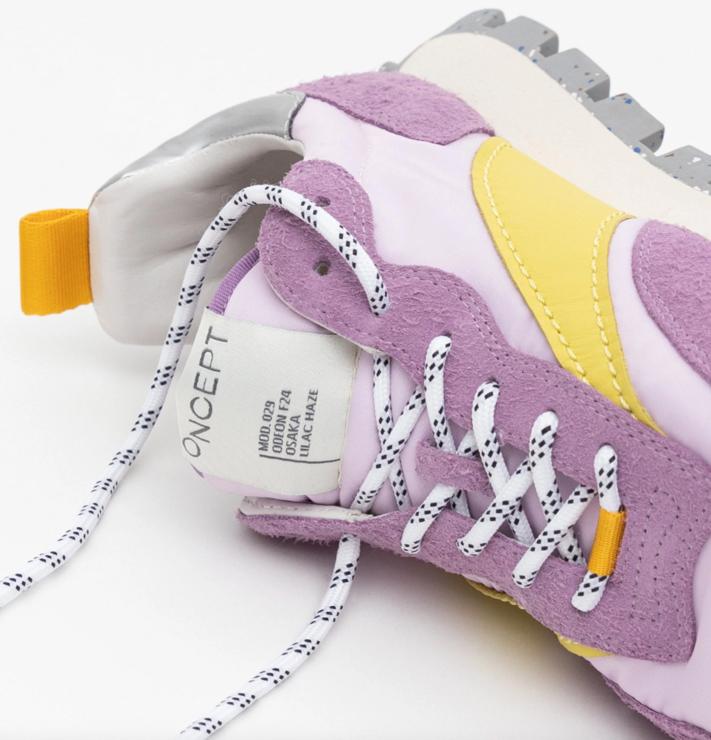 Close-up of the Osaka Lilac Haze Re-Nylon shoe highlighting its purple suede, yellow accents, and white mesh. This Osaka-inspired sneaker features black-and-white speckled laces, a rugged gray sole with a platform midsole, and orange fabric details. The tongue displays a visible white tag with text.