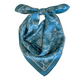 The Denim Scarf is an elegant handmade piece from Montreal, featuring a blue silk fabric adorned with intricate white floral and leaf patterns. The scarf is finished with a neatly tied bow, embodying effortless style.