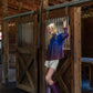 A person leaning against the open door of a rustic stable is wearing the Charlotte Top in Midnight Ombre, paired with white shorts and purple cowboy boots. The setting features string lights that contribute to a cozy atmosphere.