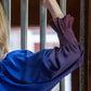 A person wearing the Midnight Ombre Charlotte Top leans an arm against vertical metal bars. The sleeve is adorned with a gathered design at the wrist. Warm, blurred lights glow in the background.