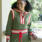 A person with curly hair smiles while leaning on a railing, dressed in their stylish Downtown Jogger - Olive Night Monogram loungewear. They stand in front of a bright, green outdoor setting, creating a vibrant scene that contrasts beautifully with the bustling city life.