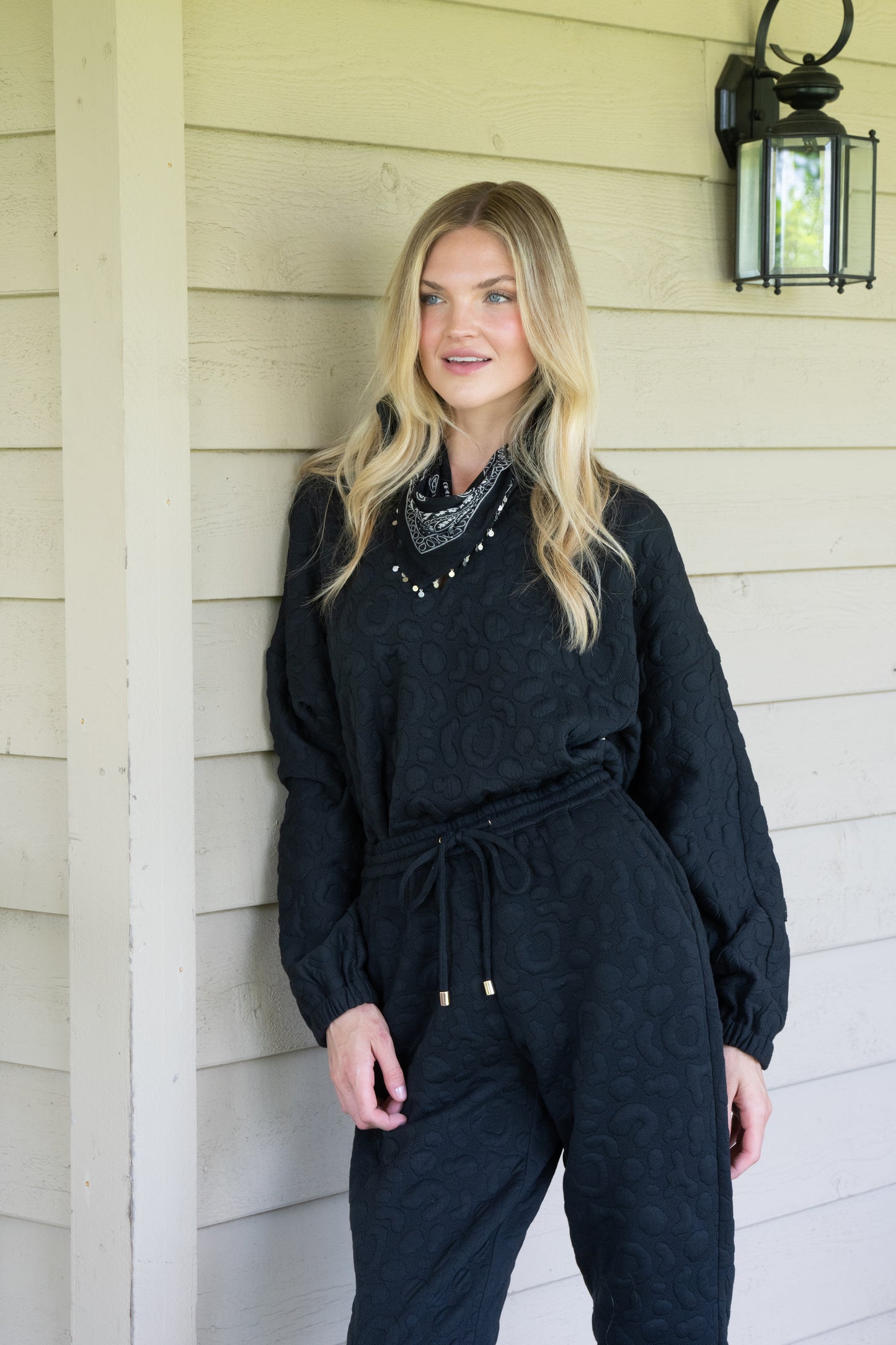 A person with long blonde hair stands against a light-colored wooden wall, donning the Poppy Pop-Over in Jet Black Cheetah and a patterned neck scarf. A black outdoor lantern is mounted on the wall above.