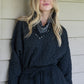 A person with long blonde hair, dressed in a black Poppy Pop-Over made from Jet Black Cheetah fabric and a patterned bandana, stands against a wood-paneled wall, illuminated by soft lighting and a nearby wall sconce.