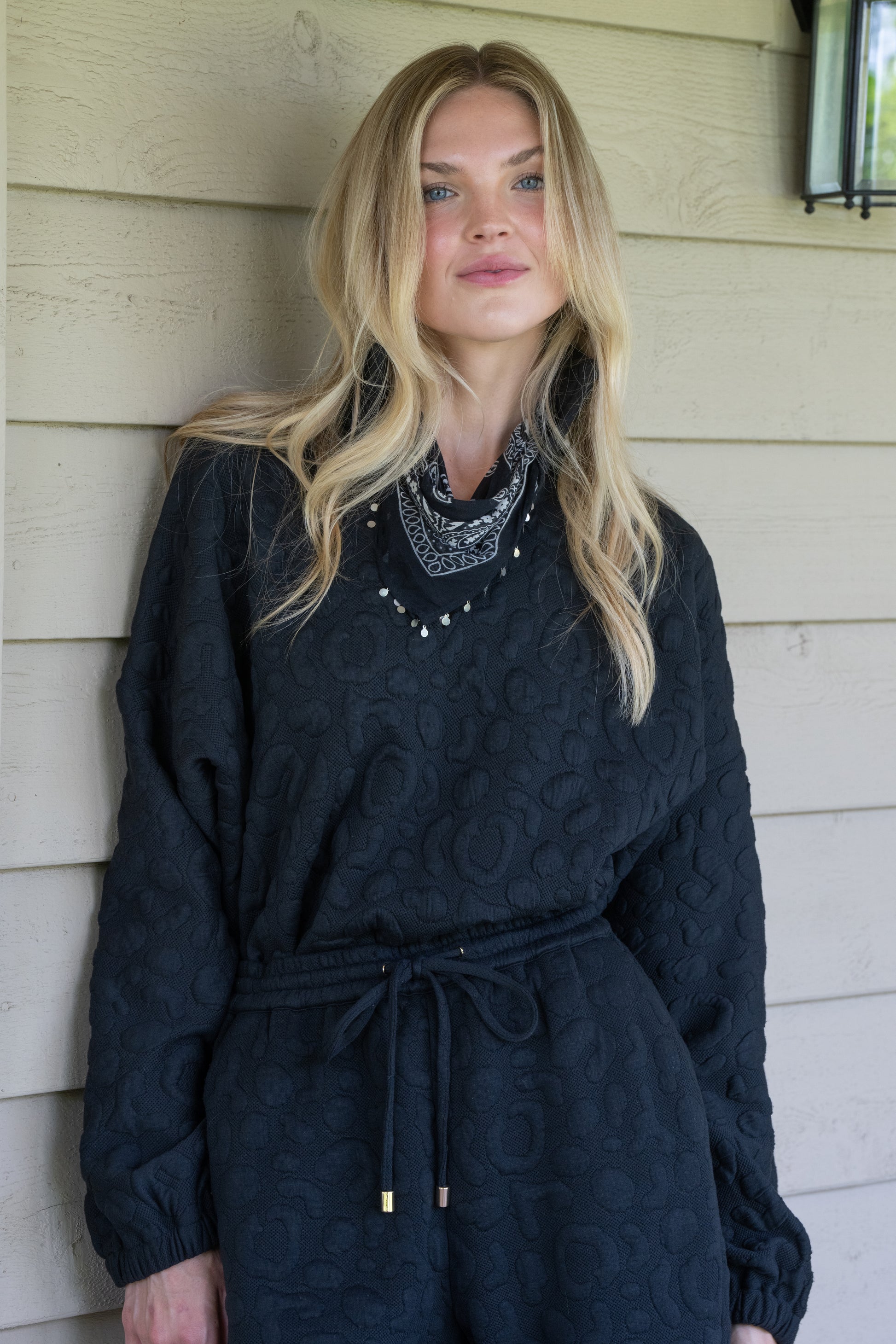 A person with long blonde hair, dressed in a black Poppy Pop-Over made from Jet Black Cheetah fabric and a patterned bandana, stands against a wood-paneled wall, illuminated by soft lighting and a nearby wall sconce.