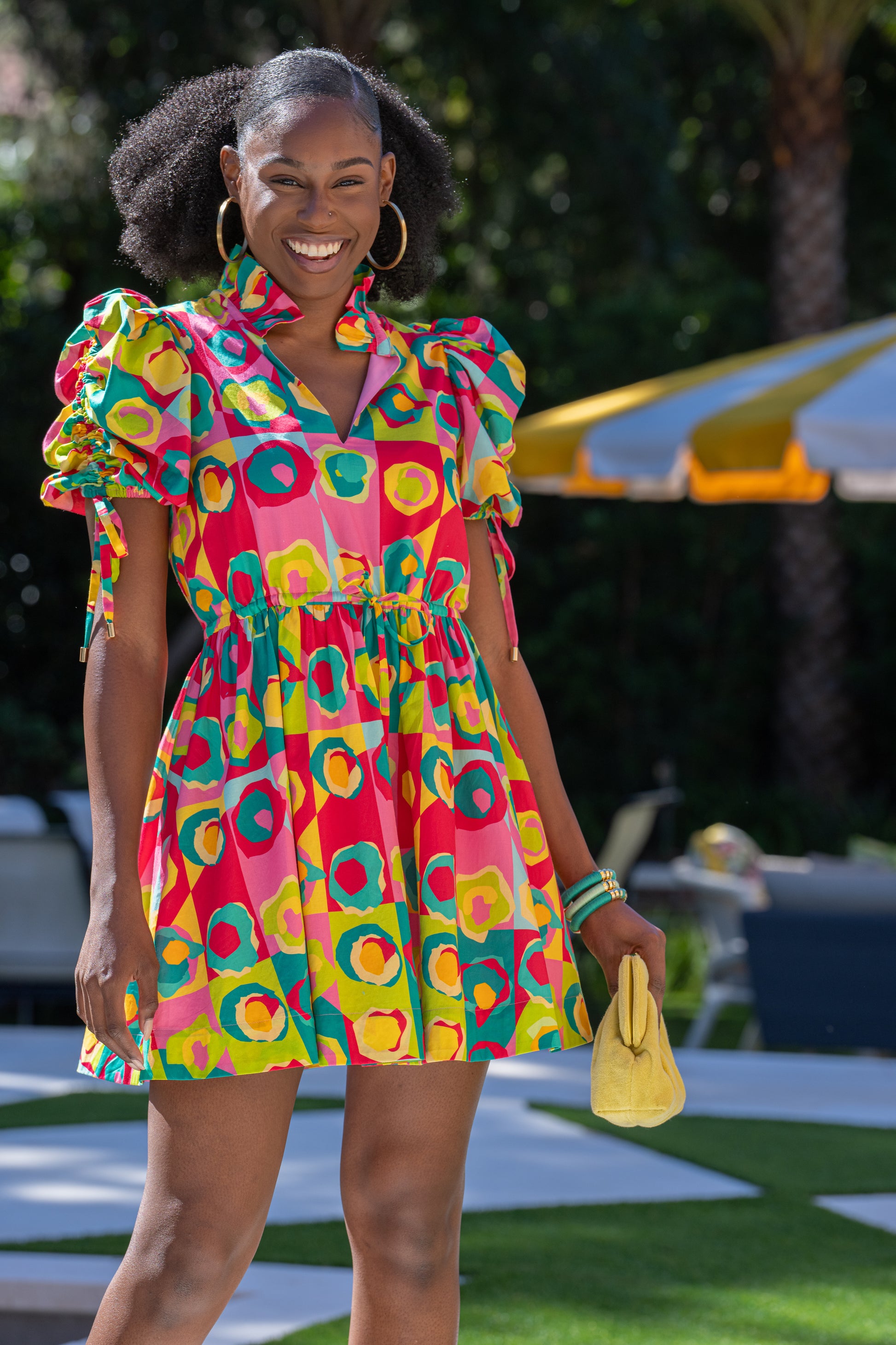 A person beams while standing outdoors in the vibrant Posey Dress - Rhett Pop, featuring a geometric pattern and puff sleeves. They hold a small yellow clutch and wear hoop earrings. The scene behind them showcases a green lawn, trees, and a yellow-striped umbrella.