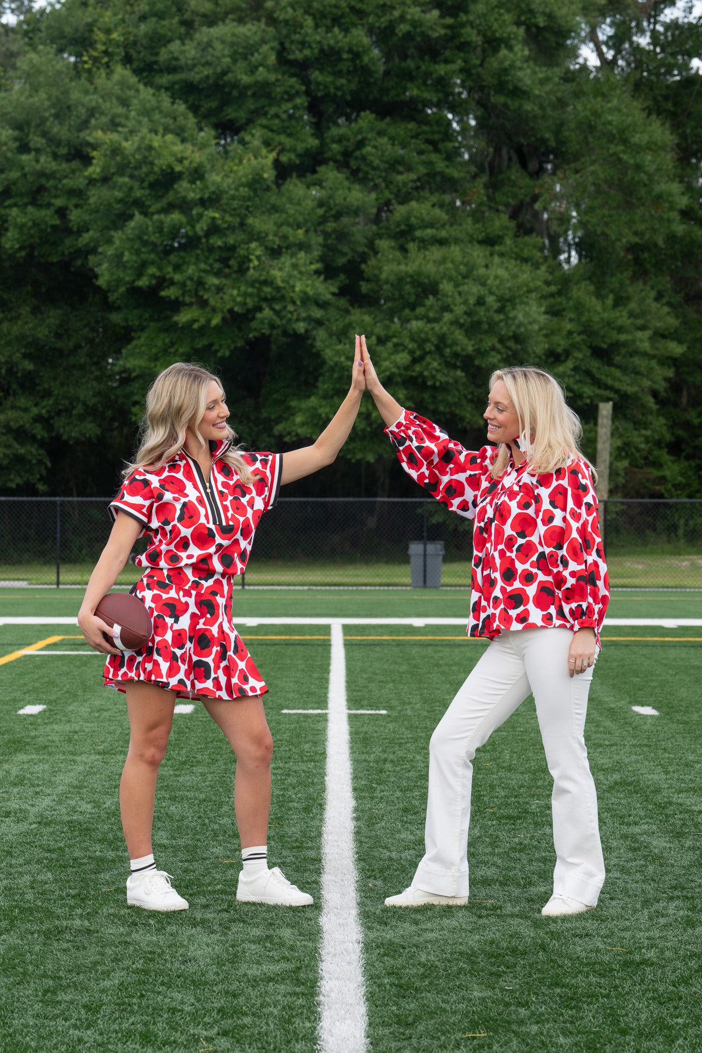 Two women, dressed in matching Stella Tops in Red Collegiate Cheetah with a signature loose fit, high-five each other on a football field. One holds a football as they smile brightly, standing on a marked line with trees providing the perfect backdrop.