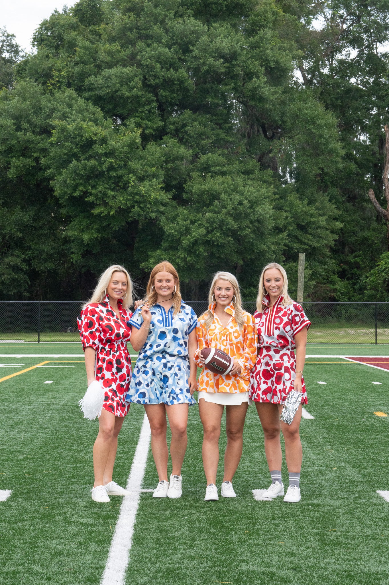 Four women stand on a football field wearing the Poppy Pullover in Burgundy Collegiate Cheetah with dolman sleeves, each holding pom-poms and a football. They are poised perfectly on the field's white line, framed by a backdrop of trees swaying gently in the breeze.