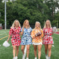 Four women, each wearing the Stella Top in Orange Collegiate Cheetah from the Emily McCarthy Collection, stroll arm in arm on a football field. Each holds a themed item: cheer pom-poms, a football, and a megaphone. They’re smiling with trees and a goalpost in the background, embodying a relaxed empire shape style.