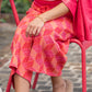 A person is seated on a red chair, wearing the Teddy Crop Trouser with a vibrant pink and orange geometric pattern, complemented by a pink jacket draped over their shoulders and beige loafers adorned with fur details. They rest their hand on their knee, effortlessly blending style with comfort in their chic viscose knit ensemble.