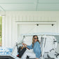 A woman wearing sunglasses and an Oxford - Denim Chambray blouse with signature gold buttons is seated in a white and blue floral-patterned golf cart parked inside a white garage. She smiles while holding the steering wheel. The garage features white wooden doors and a pale blue ceiling.