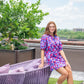 A person in a vibrant Poppy Dress - Bauhaus with loose dolman sleeves sits on a purple outdoor chair on a rooftop patio. They're wearing white boots and smiling, surrounded by green plants with a view of the cityscape under a cloudy sky.