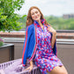 A person with long red hair poses outdoors, sitting on the back of a purple chair. They have the Clara Cardigan in Bauhaus Stripe draped over their shoulders and a colorful geometric-patterned dress, surrounded by greenery and a building in the background.