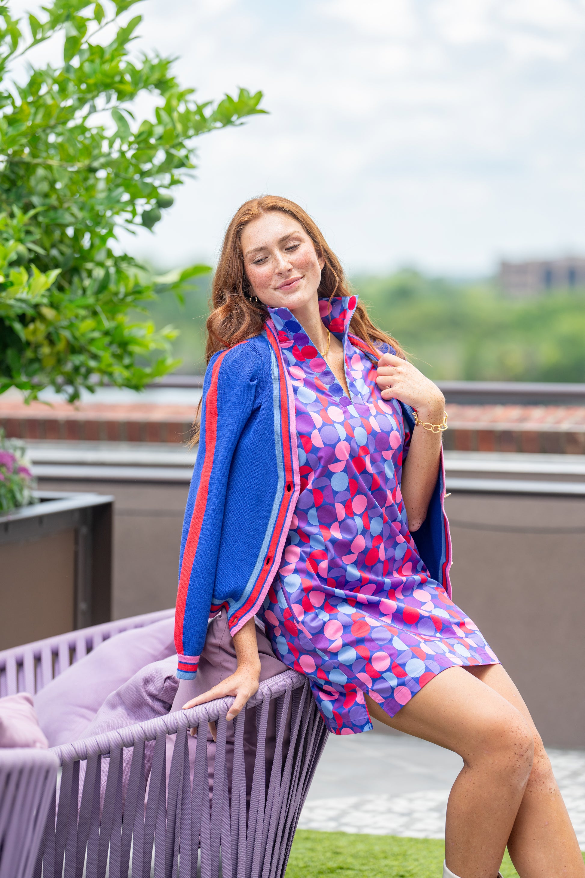 A person with long red hair poses outdoors, sitting on the back of a purple chair. They have the Clara Cardigan in Bauhaus Stripe draped over their shoulders and a colorful geometric-patterned dress, surrounded by greenery and a building in the background.