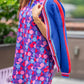 A woman stands outdoors in a vibrant, geometric-patterned dress with puff sleeves, splashed in shades of pink, purple, and blue. Draped over her shoulders is the Clara Cardigan in Bauhaus Stripe with gold button closures. Her look is completed with a gold bracelet on her wrist.