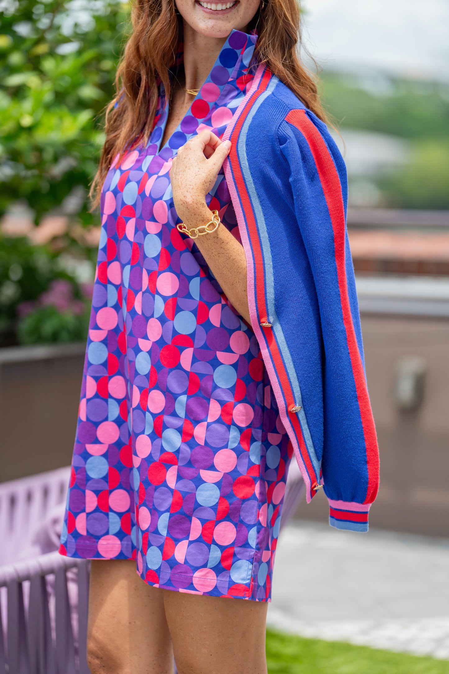 A woman stands outdoors in a vibrant, geometric-patterned dress with puff sleeves, splashed in shades of pink, purple, and blue. Draped over her shoulders is the Clara Cardigan in Bauhaus Stripe with gold button closures. Her look is completed with a gold bracelet on her wrist.