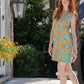 A woman with long red hair stands on a sunlit outdoor patio, wearing the Emily Shift Dress - Poolside by Emily McCarthy Clothing. Bright orange flowers bloom nearby, and the lush green foliage offers a perfect backdrop. A vintage-style wall lantern adds charm to the scene.