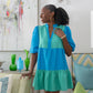 In a chic living room, a woman wearing the Frankie Dress in Lakeside Colorblock—featuring vibrant blue and green hues—smiles while touching a chair. She is surrounded by colorful decor, including green glass vases and an abstract painting.