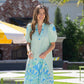 A woman wearing the Hampton Dress - Cordoba with puff sleeves, showcasing blue patterns, stands on a patio. She smiles with trees and a brick structure in the background. The setting seems sunny and relaxed, ideal for highlighting the dress's versatile styling.