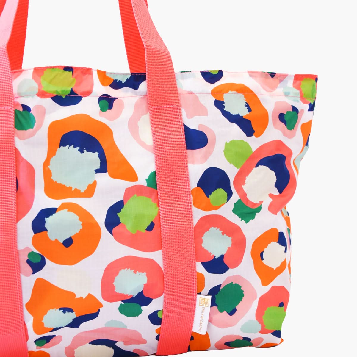 This Travel Tote - Multi Spot Cheetah showcases vivid pink handles and a striking abstract pattern with splashes of orange, blue, green, and pink set against a white backdrop. As part of the Emily McCarthy Collection, it serves as an exceptional travel tote for individuals who appreciate vibrant style.