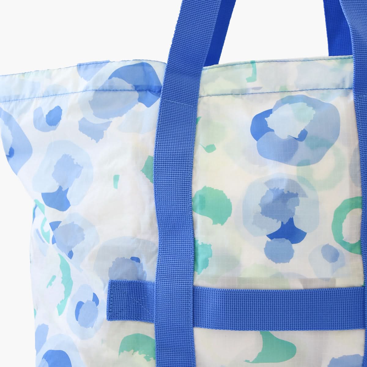 A close-up of the Seaglass Spot Cheetah travel tote by Emily McCarthy highlights its blue and green abstract circular designs on a white background. The tote is equipped with sturdy blue straps and provides a partial view of the top and side panels.