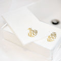 A stack of 1-Letter Monogrammed Guest Towels with an ornate gold design is displayed in a clear acrylic holder on a white surface, adding elegance to any setting.