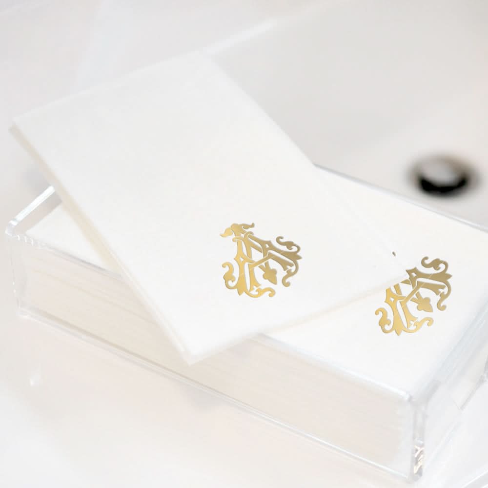 A stack of 1-Letter Monogrammed Guest Towels with an ornate gold design is displayed in a clear acrylic holder on a white surface, adding elegance to any setting.