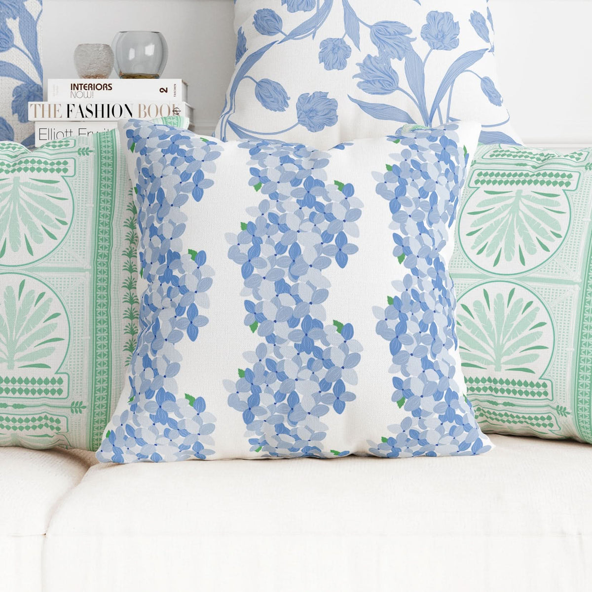 A decorative couch features five pillows, with the central one adorned in bold botanical patterns from the "Fabric by the Yard - Hydrangea Stripe" collection, showcasing blue and green floral designs. Made from a cotton and linen blend, it is accompanied by two light green geometric patterned pillows and two hand-drawn blue floral motif pillows, all on a crisp white background.