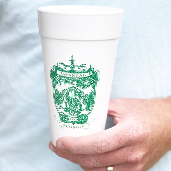 A person in a light blue shirt holds a white cup from the Savannah Crest Foam Cup Set, showcasing the words Savannah and Georgia, with a ship and the date 1733, adding historic charm to this drinkware.