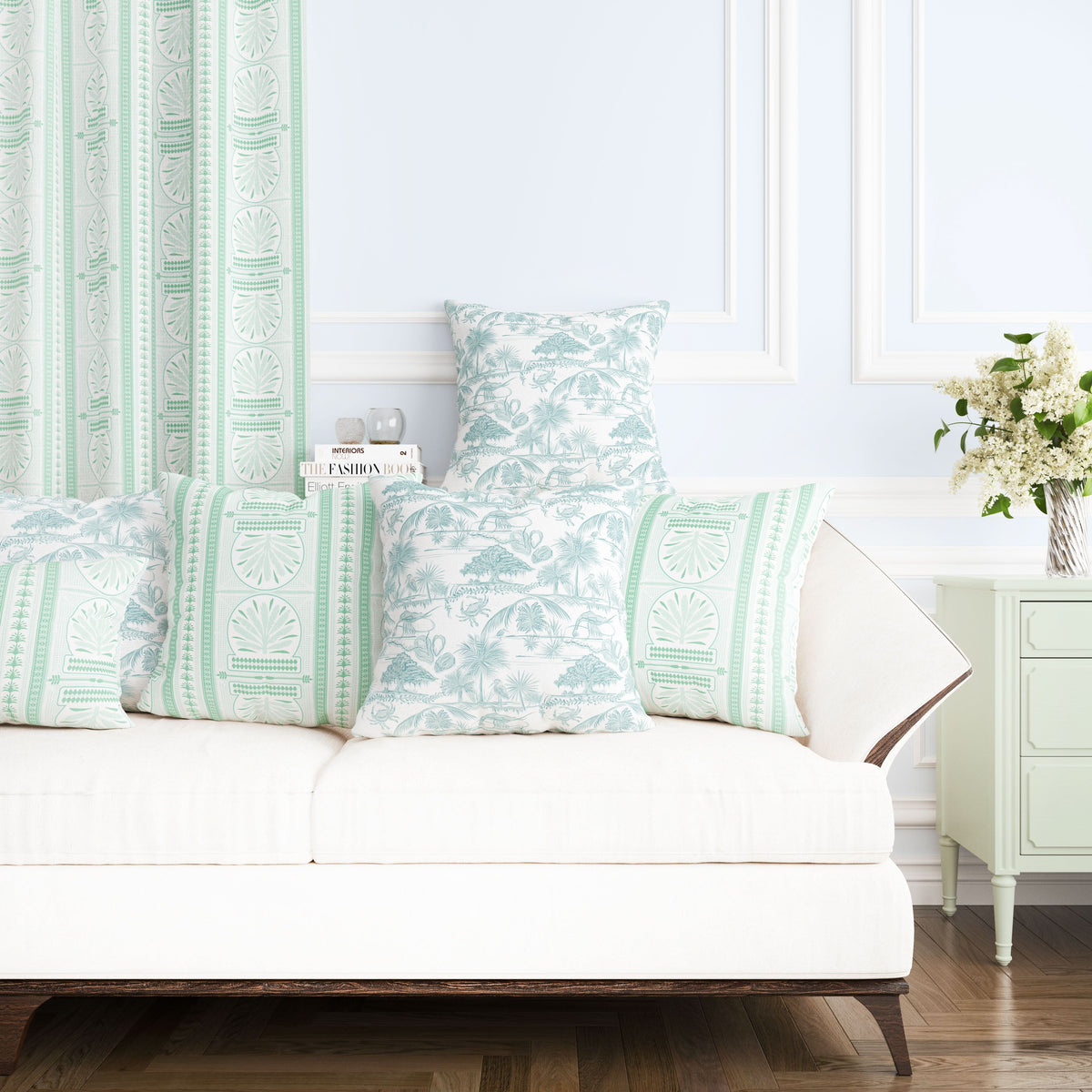 A light and airy living room showcases a white sofa embellished with mint green and white patterned pillows. A small white table supports a potted plant with delicate blossoms. The walls are light blue, complemented by modern deco Royal Palm print curtains from Fabric by the Yard, which perfectly coordinate with the pillows.