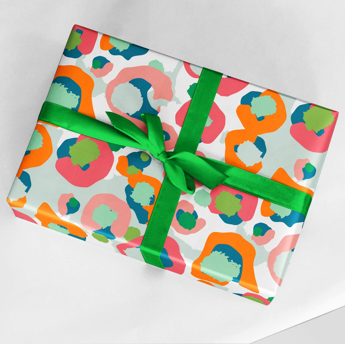 A gift wrapped in the vibrant Giftwrap - Multi Spot Cheetah paper, featuring an abstract cheetah pattern with orange, pink, blue, and green shapes. It is tied with a bright green ribbon that forms a bow on top and is placed against a white background.