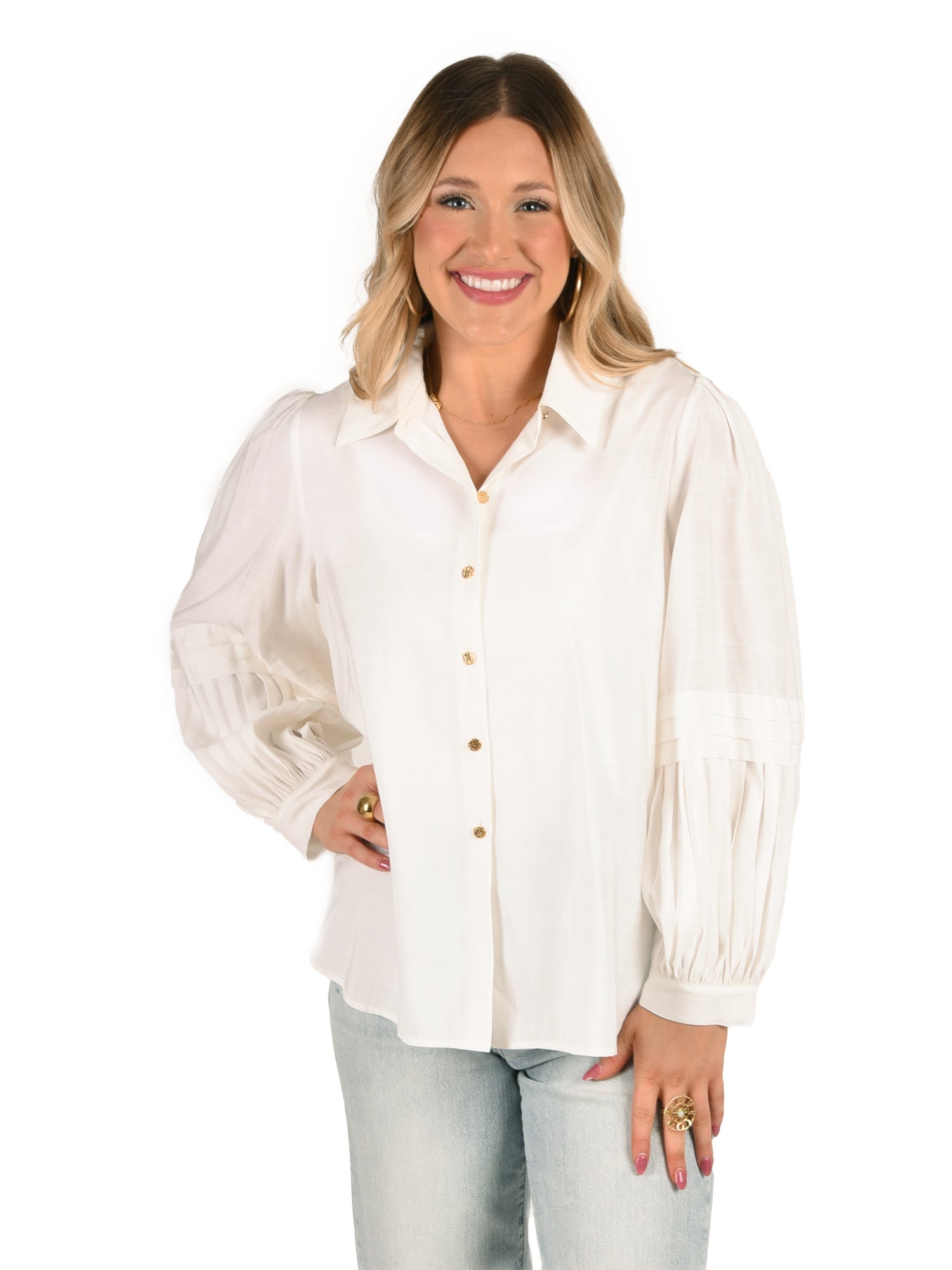 A woman smiles while wearing the chic Oxford - Linen White shirt, featuring signature gold buttons and pleated balloon sleeves, perfectly paired with light blue jeans. Her shoulder-length brown hair cascades softly as she stands against a plain white background.