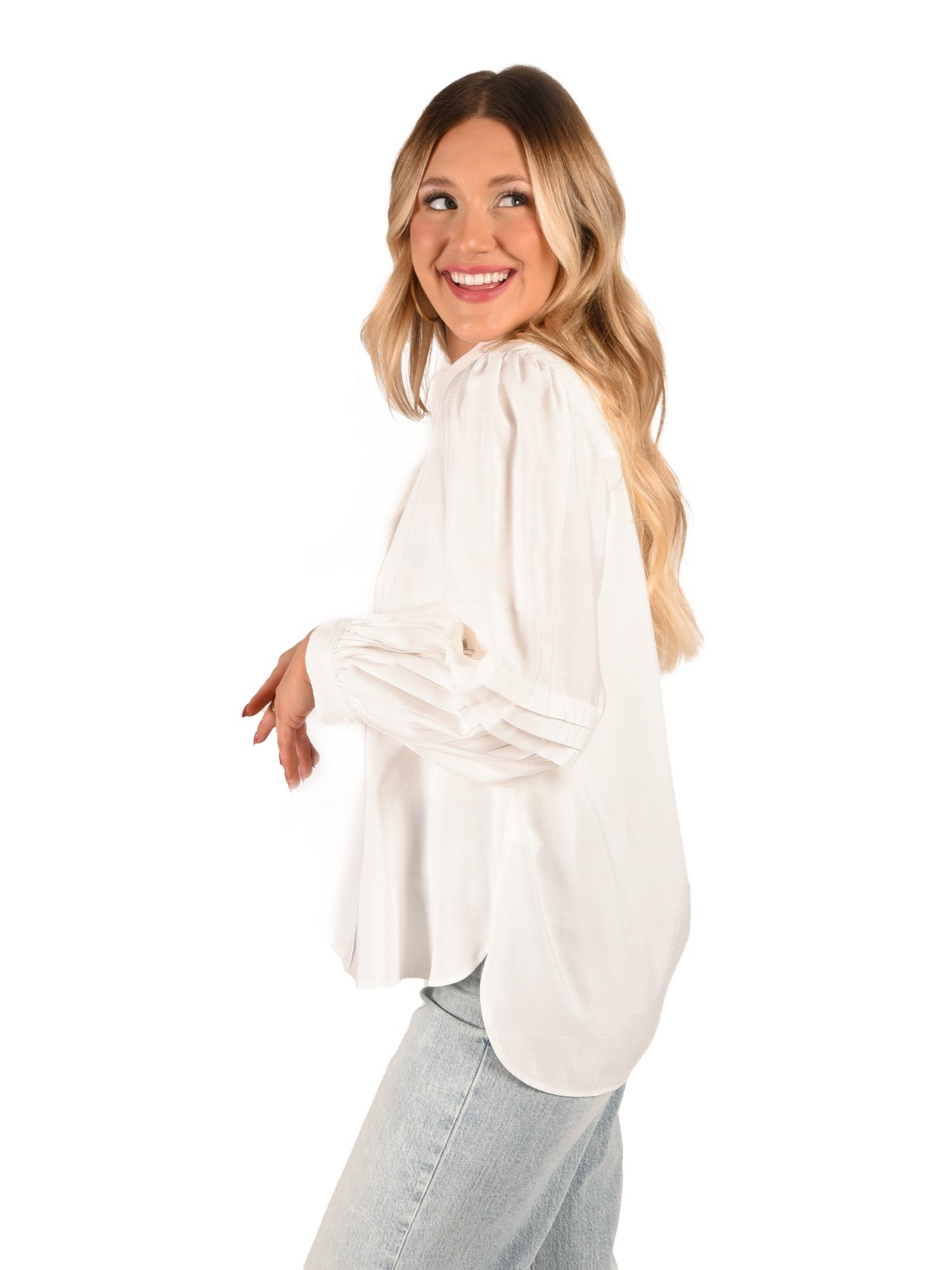 A woman with long, wavy hair smiles while facing slightly to the side. She is wearing the Oxford - Linen White blouse with pleated balloon sleeves and light blue jeans, accentuated by signature gold buttons against a plain white background.