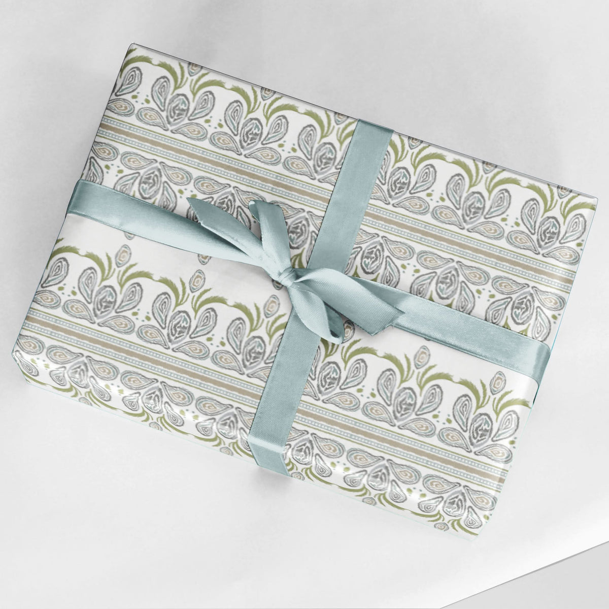 A gift wrapped in Oyster Ikat patterned paper, featuring green and gray leafy designs, is tied with a light blue ribbon and placed on a white surface.
