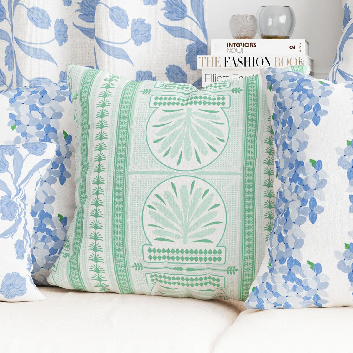 A cozy living room scene highlights a modern decorative statement with a pillow featuring green botanical patterns, surrounded by blue floral pillows. Behind them, a stack of interior design books and a vase beautifies the white couch. The backdrop features floral curtains made from Fabric by the Yard - Royal Palm, crafted in a Cotton & Linen blend.