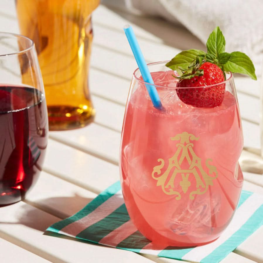 A glass of pink iced drink with a blue straw, topped with strawberry and mint, rests beside 1-Letter govino® Stemless Shatterproof Wine glasses on a striped napkin atop a sunlit white wooden table.