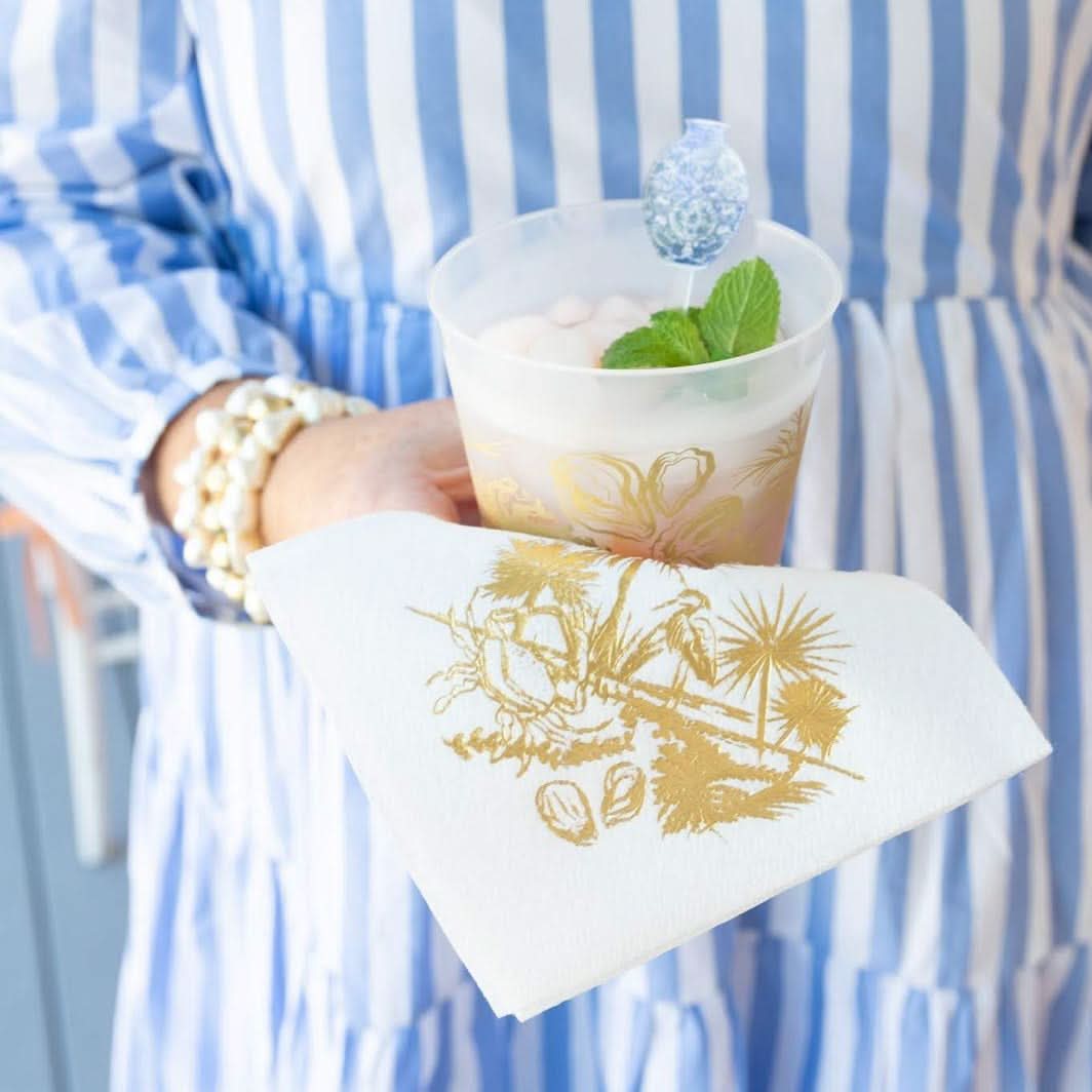 A person in a blue and white striped dress holds a mint-garnished cup, resting it on Low Country Cocktail Napkins - White & Gold. A beaded bracelet on their wrist complements the charming low country design elements.