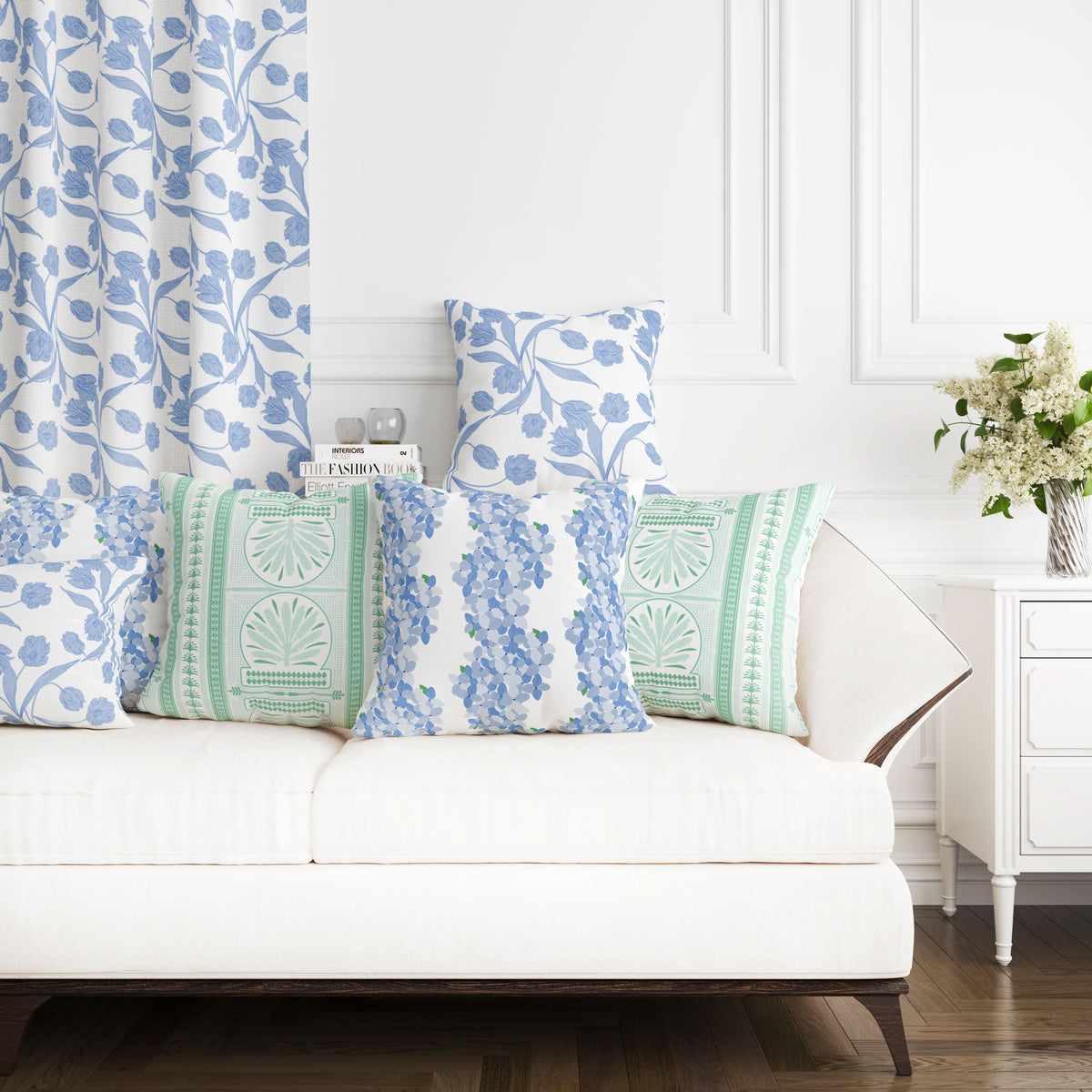 A white sofa adorned with cushions featuring a blue floral and green patterned design, crafted from a Cotton & Linen blend, is positioned against a wall with white paneling. Complementing the scene is the bold botanicals curtain made from Fabric by the Yard - Hydrangea Stripe, alongside a white side table topped with a bouquet of flowers to complete the cozy interior setting.