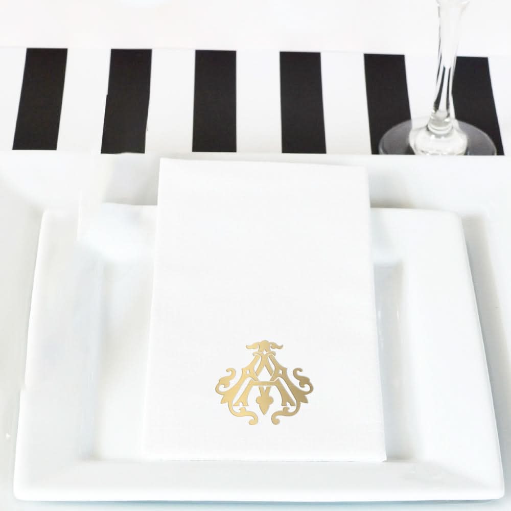 A white napkin with a gold monogram sits on a white plate. In the background are a black-and-white striped table runner, part of a wine glass, and hints of elegant 1-Letter Monogrammed Guest Towels.